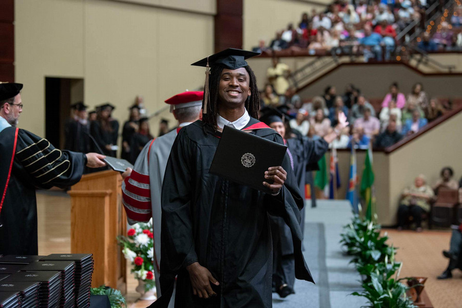 A smiling graduate in a cap and gown smiles while holding up his diploma