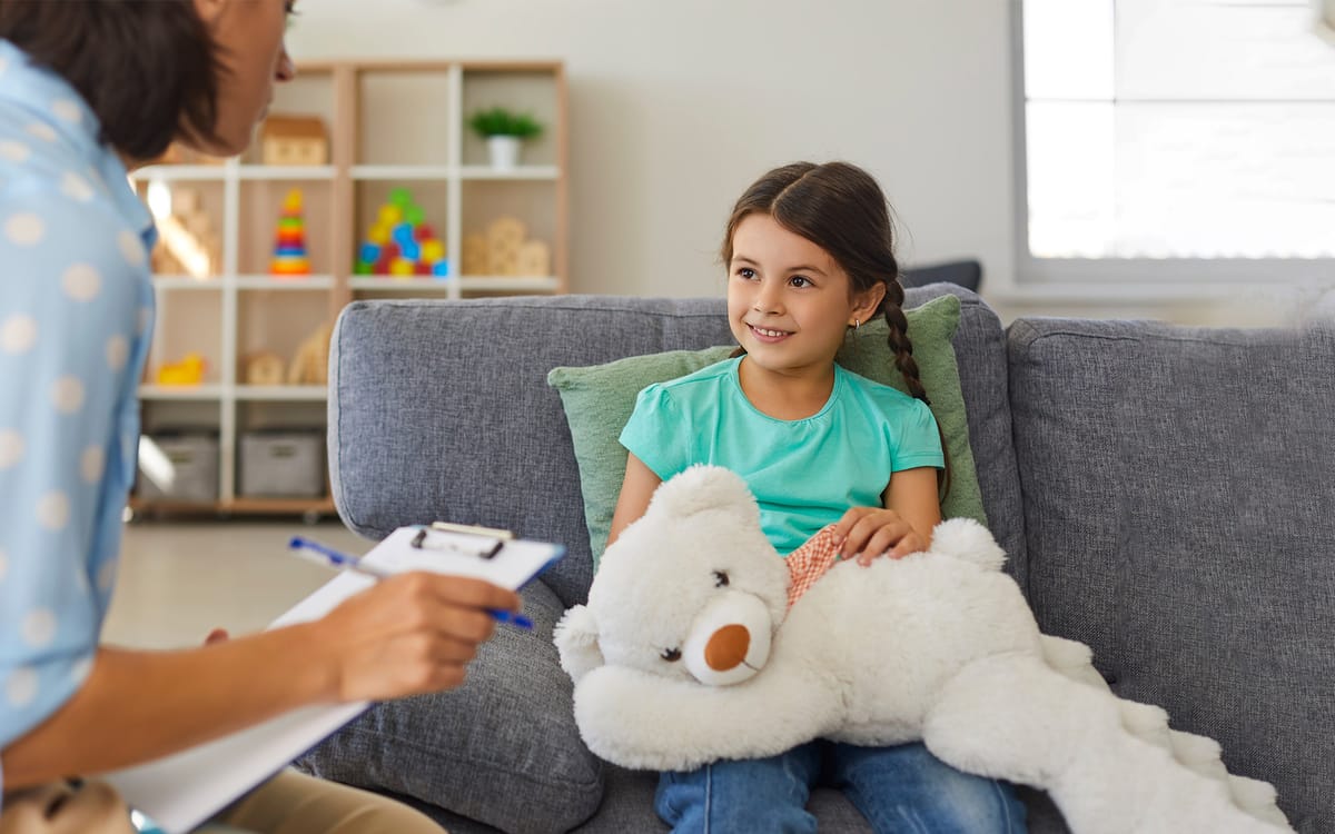 Little girl with an oversized teddy bear in her lap sitting on couch talking to counselor