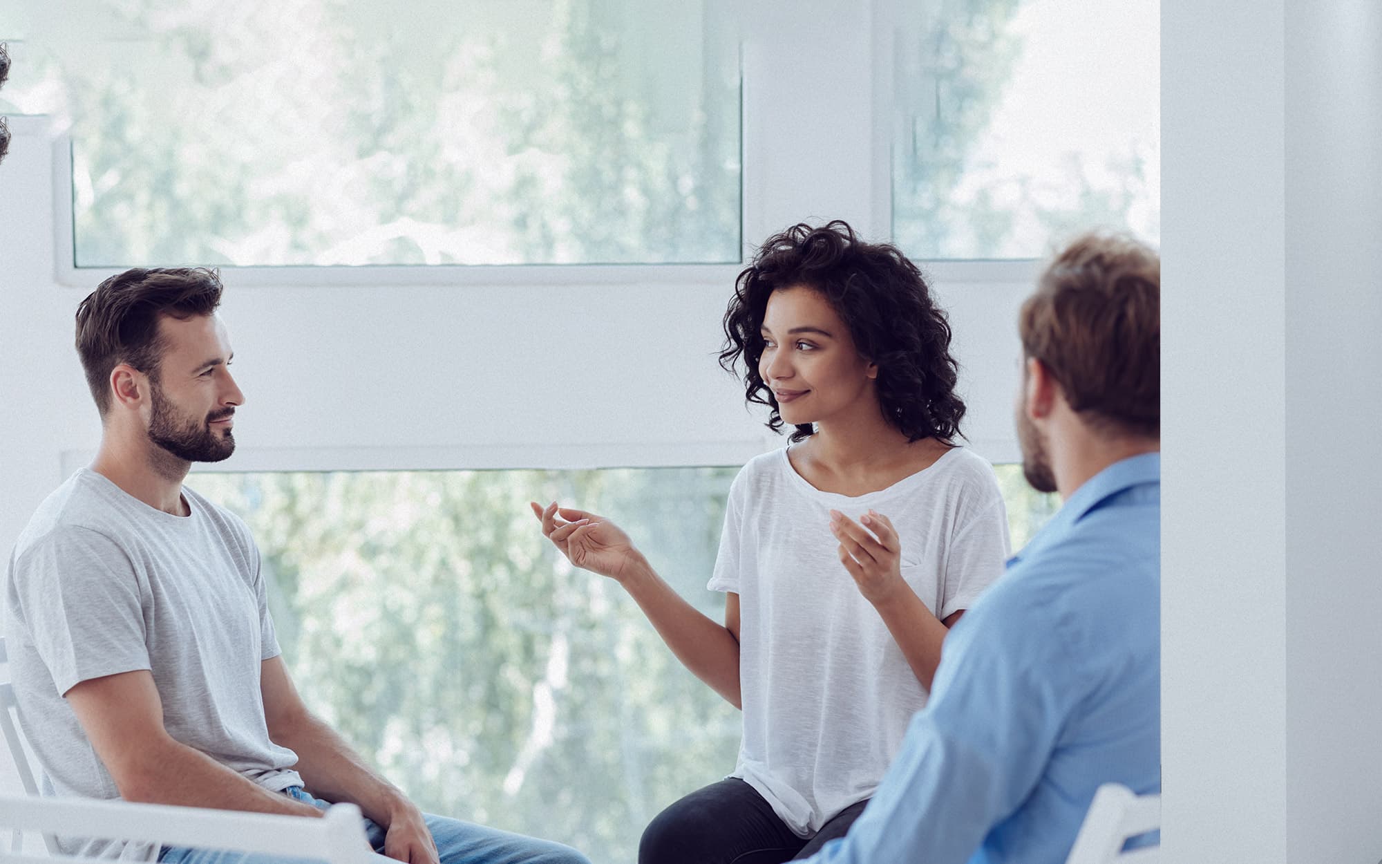 A young woman speaking and gesturing while leading a small group discussion in a bright, modern room.