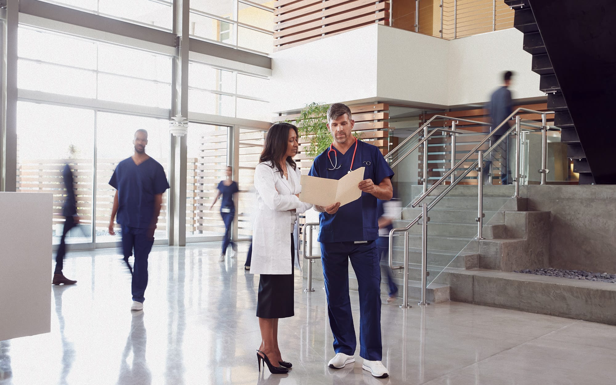 Two healthcare professionals, a woman in a white lab coat and a man in blue scrubs with a stethoscope, reviewing a patient chart together in a modern hospital lobby.