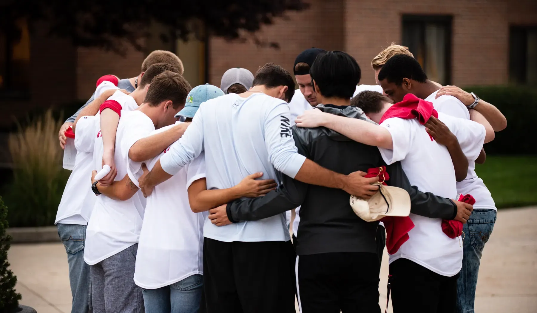 A group of young men standing in a close-knit circle with arms around each other in an outdoor setting, heads bowed in prayer.