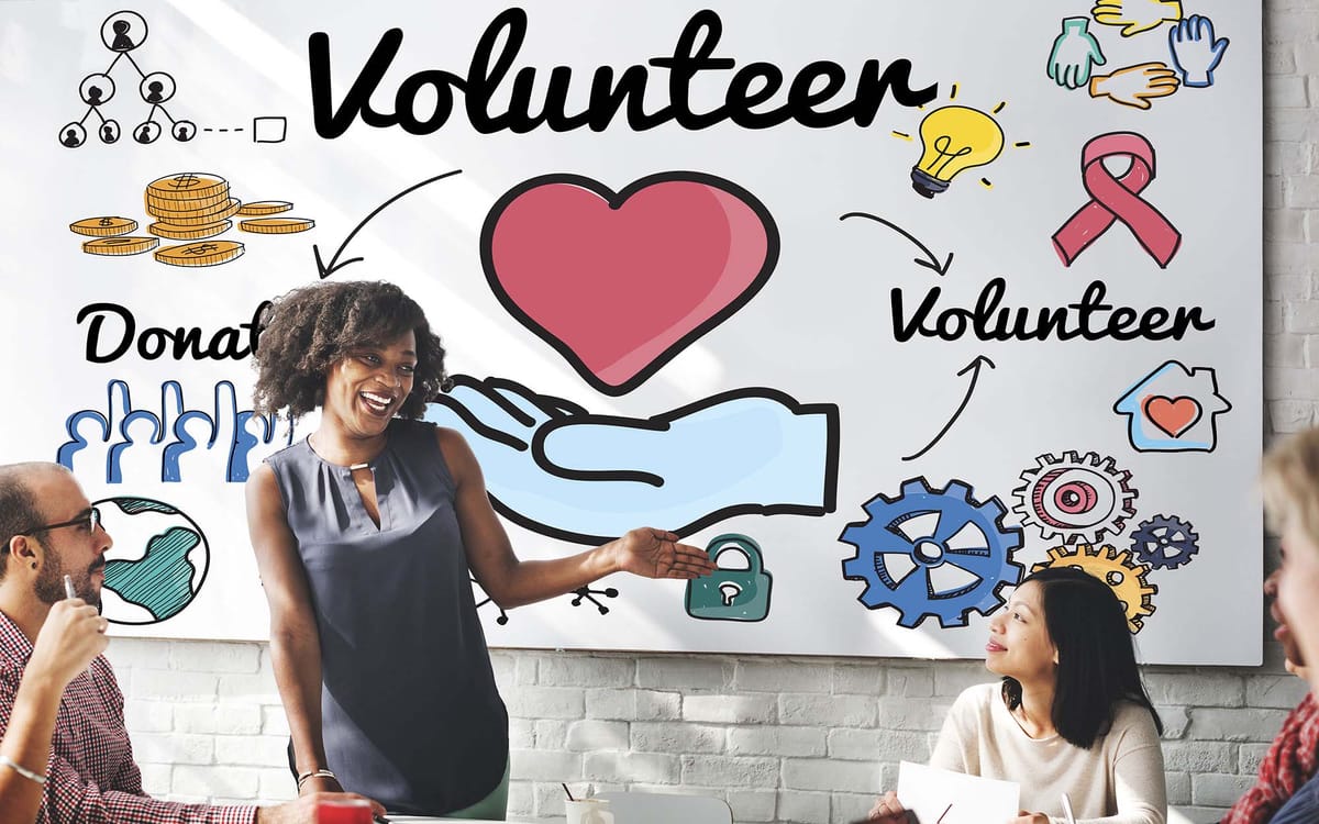 A smiling woman stands and gestures toward a large illustrated board behind her, which includes colorful graphics of a heart, hands, coins, gears, and the words "Volunteer" and "Donate"