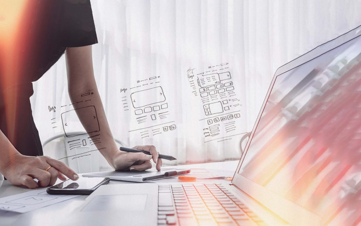 A person standing at a desk working with a tablet and stylus, referencing wireframe sketches for a digital user interface design