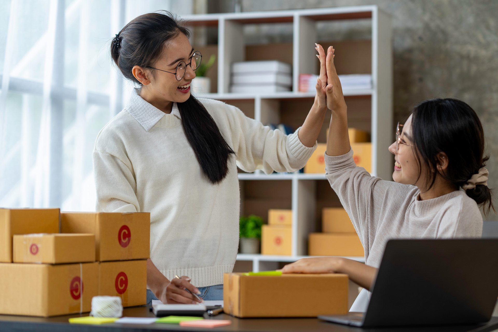 2 women high-fiving amidst boxes for small business