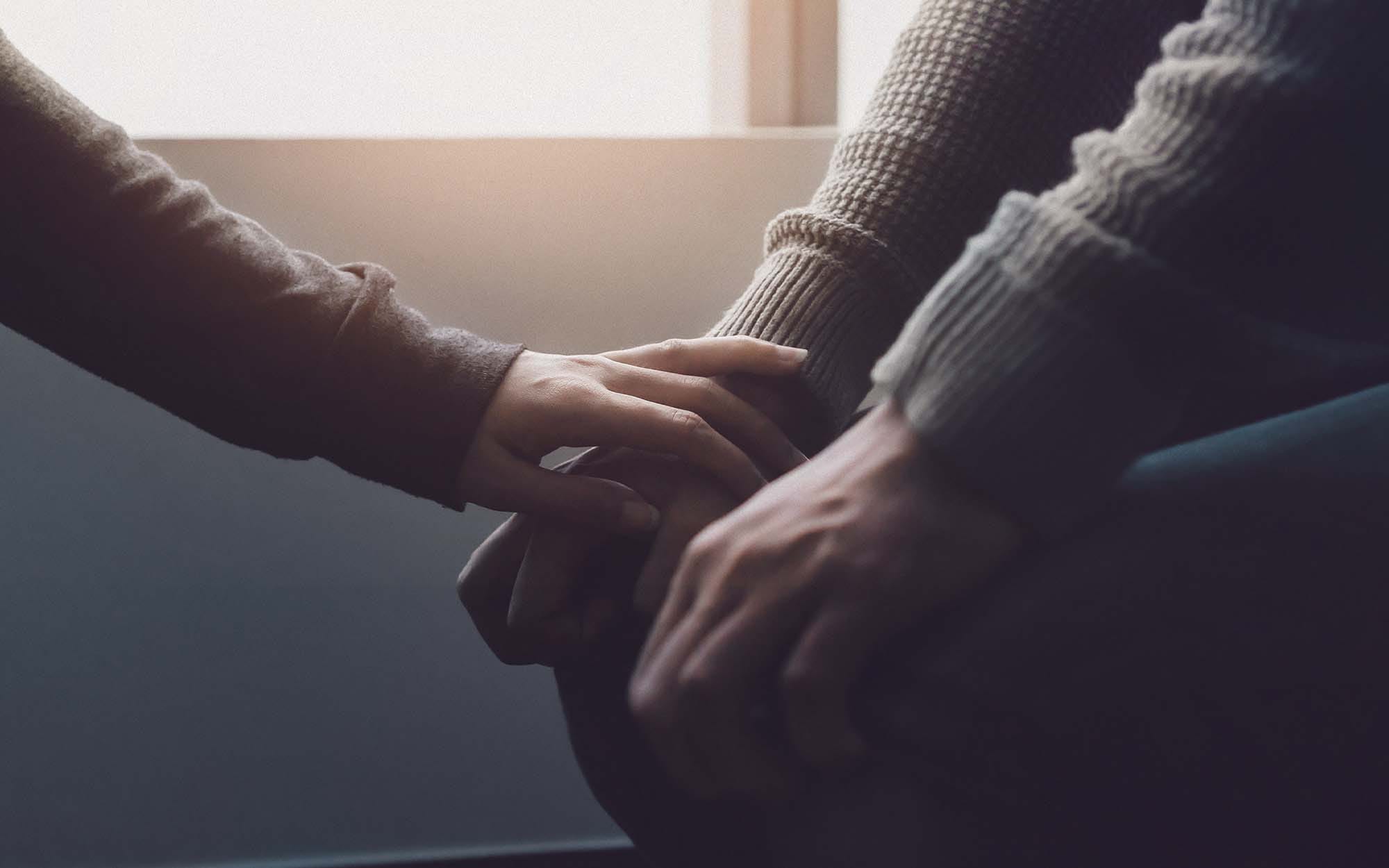 Close-up of one person gently placing a hand on another person's clasped hands in a comforting gesture