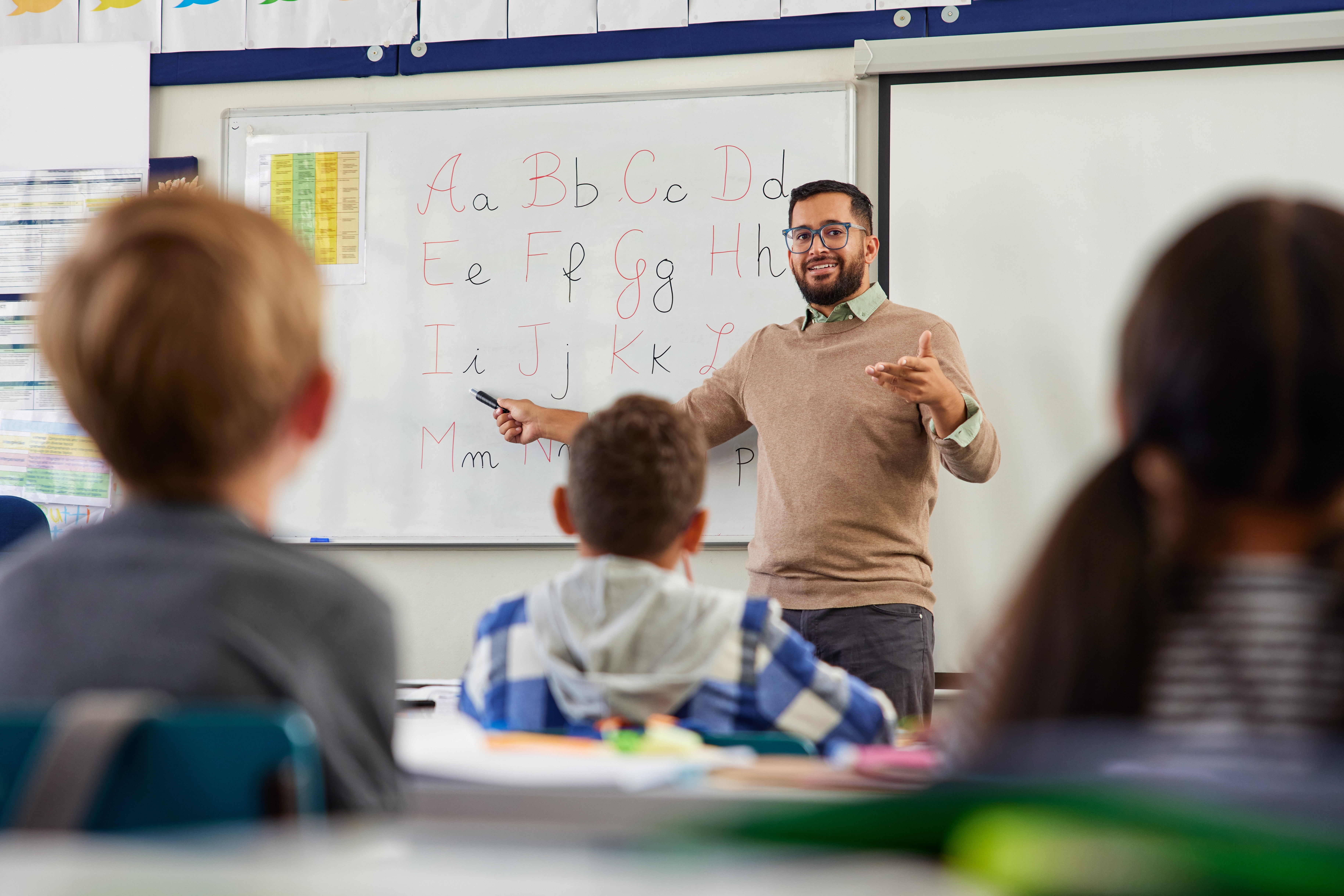 Smiling young male teacher helping students learn alphabet in first grader classroom. 