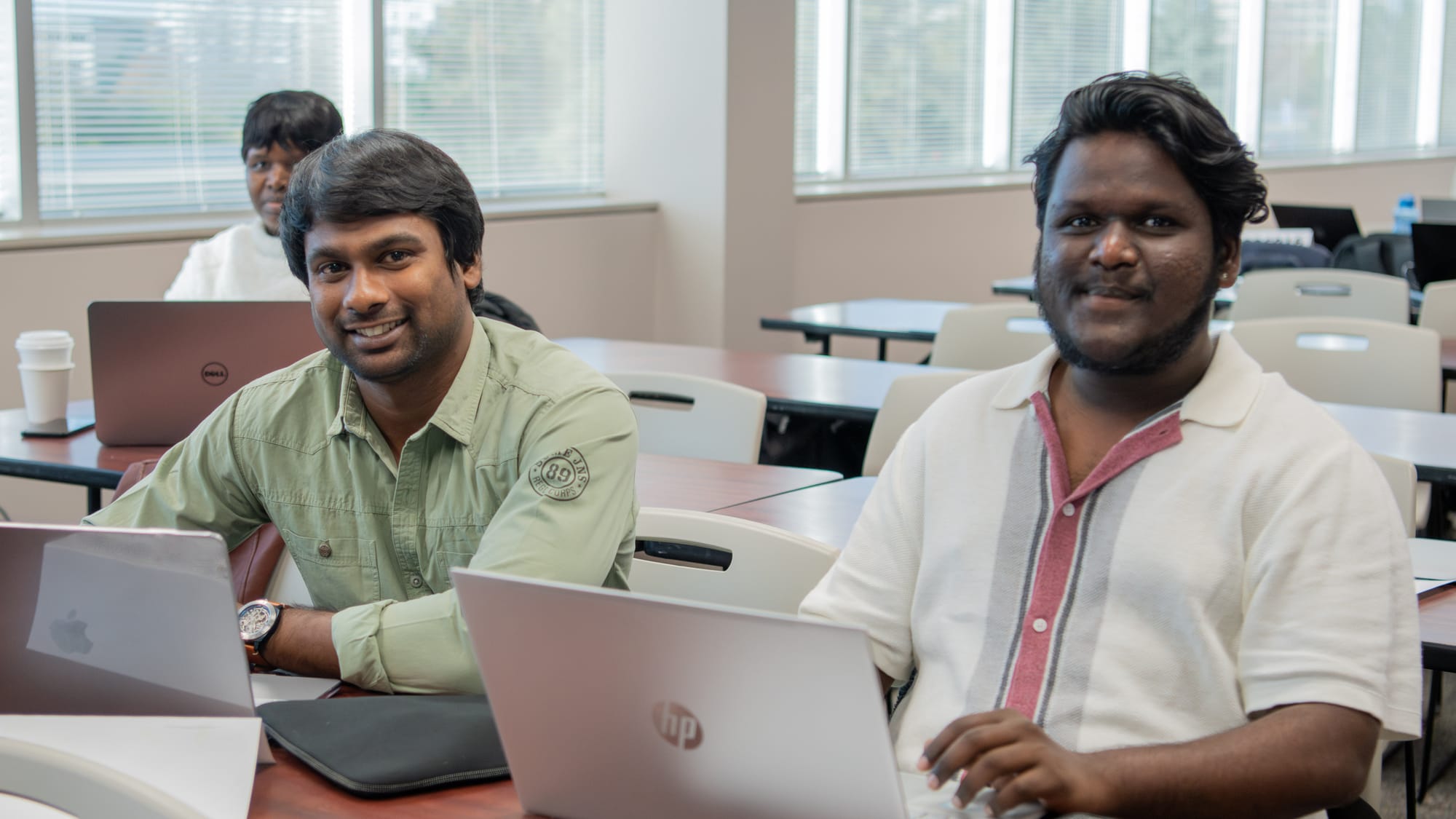 Two international students sitting at a table in a classroom, doing homework on their laptops
