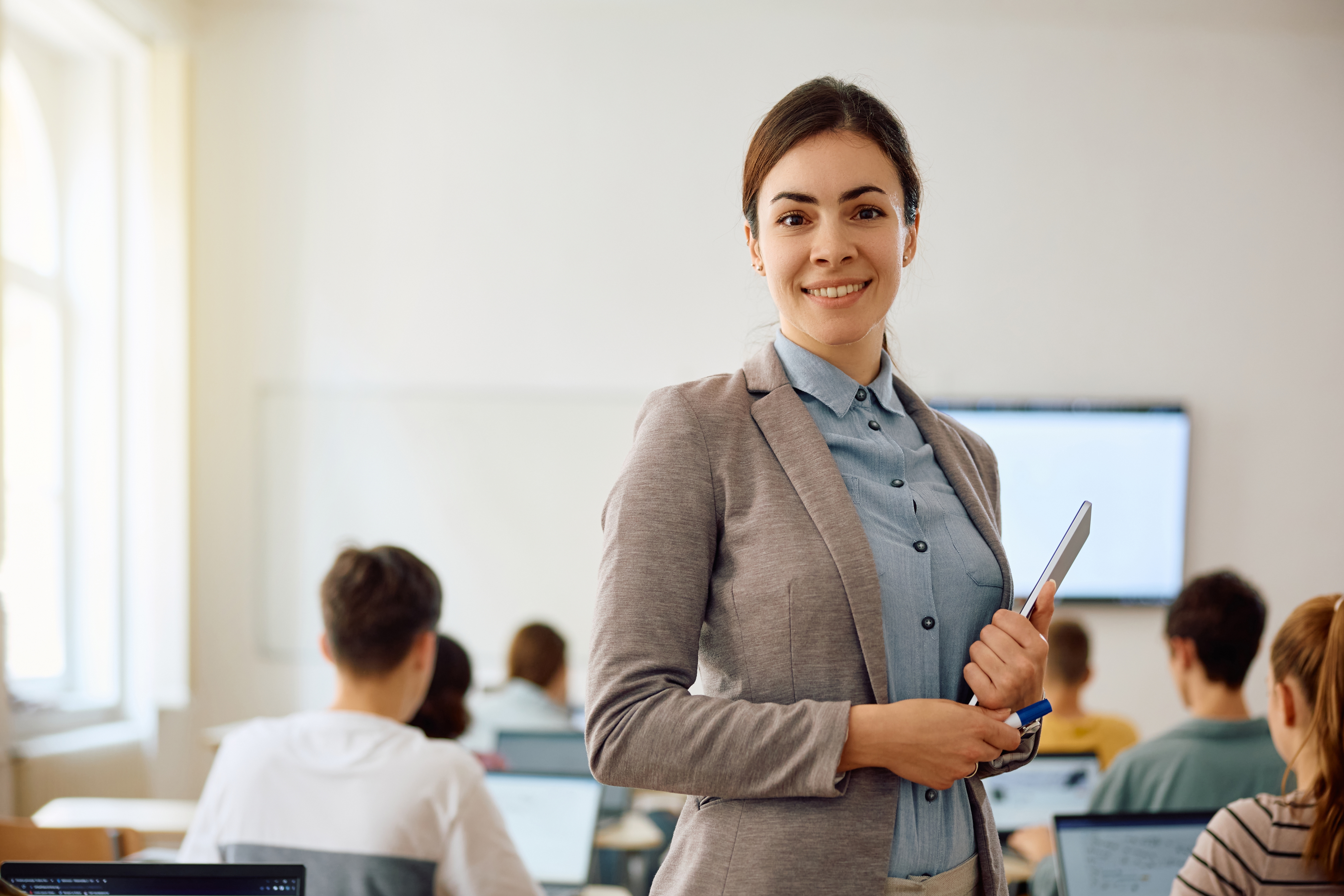 Smiling computer science teacher in the classroom and looking at camera. Her students are in the background.