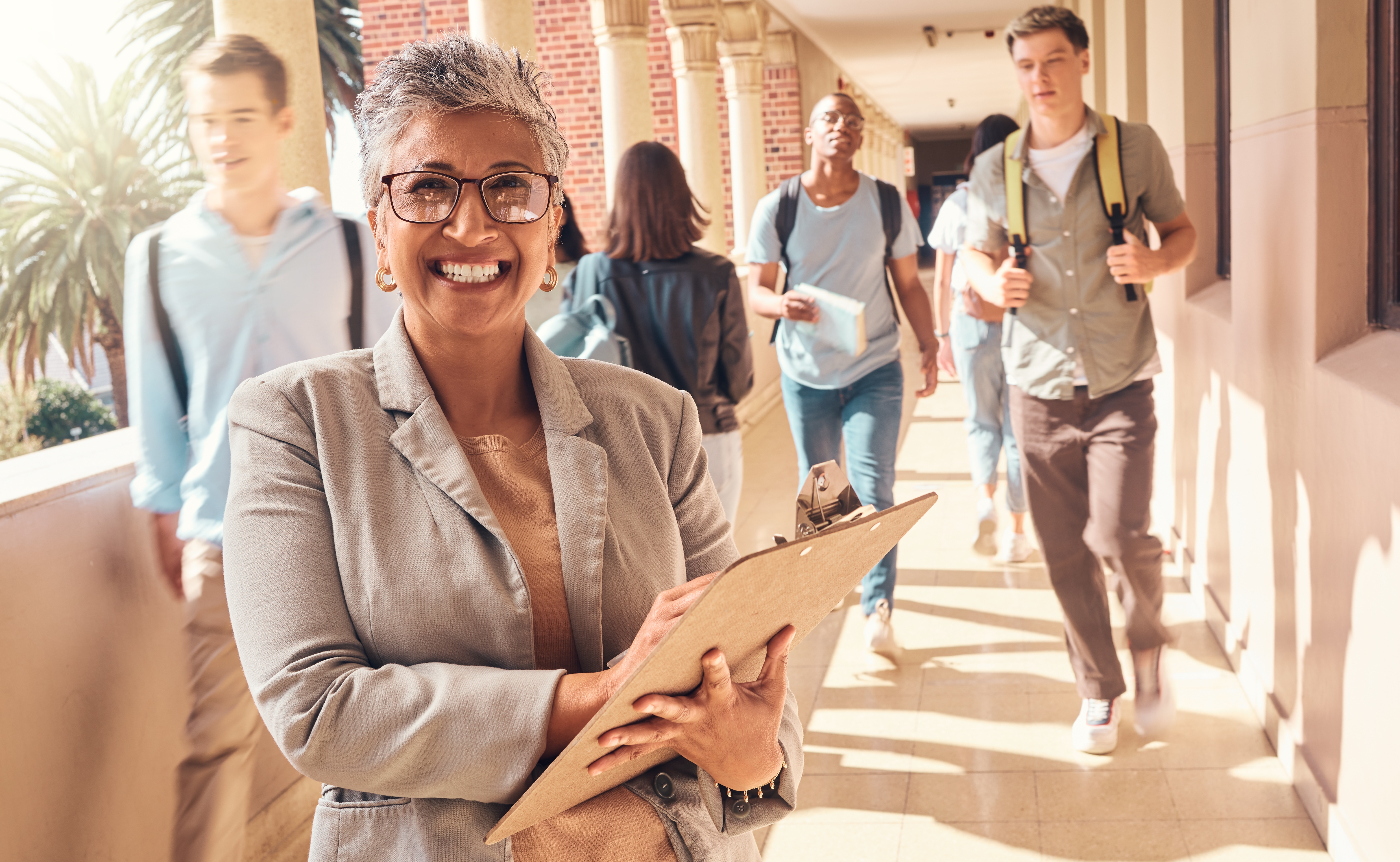 Principal smiling in front of camera while holding a clipboard. High school students are walking in the hallway behind her