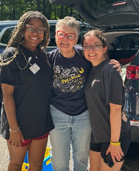 Three women smile with their arms around each other outdoors at CHAMP Camp, including two young camp volunteers and an older woman