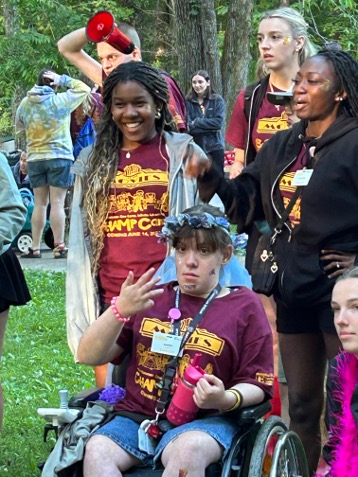 CHAMP Camp participants and volunteers gather outdoors in a wooded area, including a camper using a wheelchair surrounded by fellow campers and counselors.