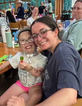 An IWU student volunteer smiles alongside a young CHAMP Camp camper with a tracheostomy while sitting together at a table indoors.