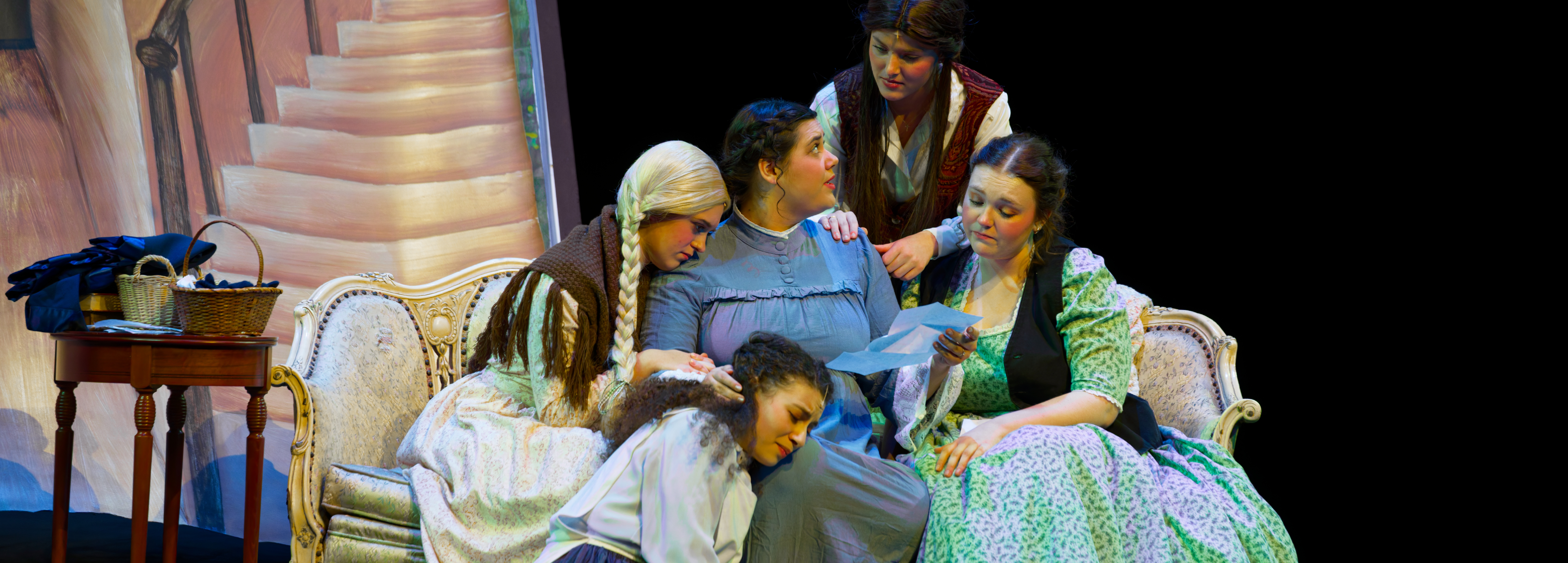 Students perform a scene from Little Women: The Broadway Musical at IWU, with the March sisters gathered closely on a couch reading a letter in a warmly lit stage setting.